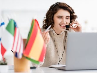 overjoyed language teacher in headset holding pen and talking near laptop and international flags on blurred foreground
